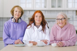 Three smiling older women in a kitchen holding coffee cups.